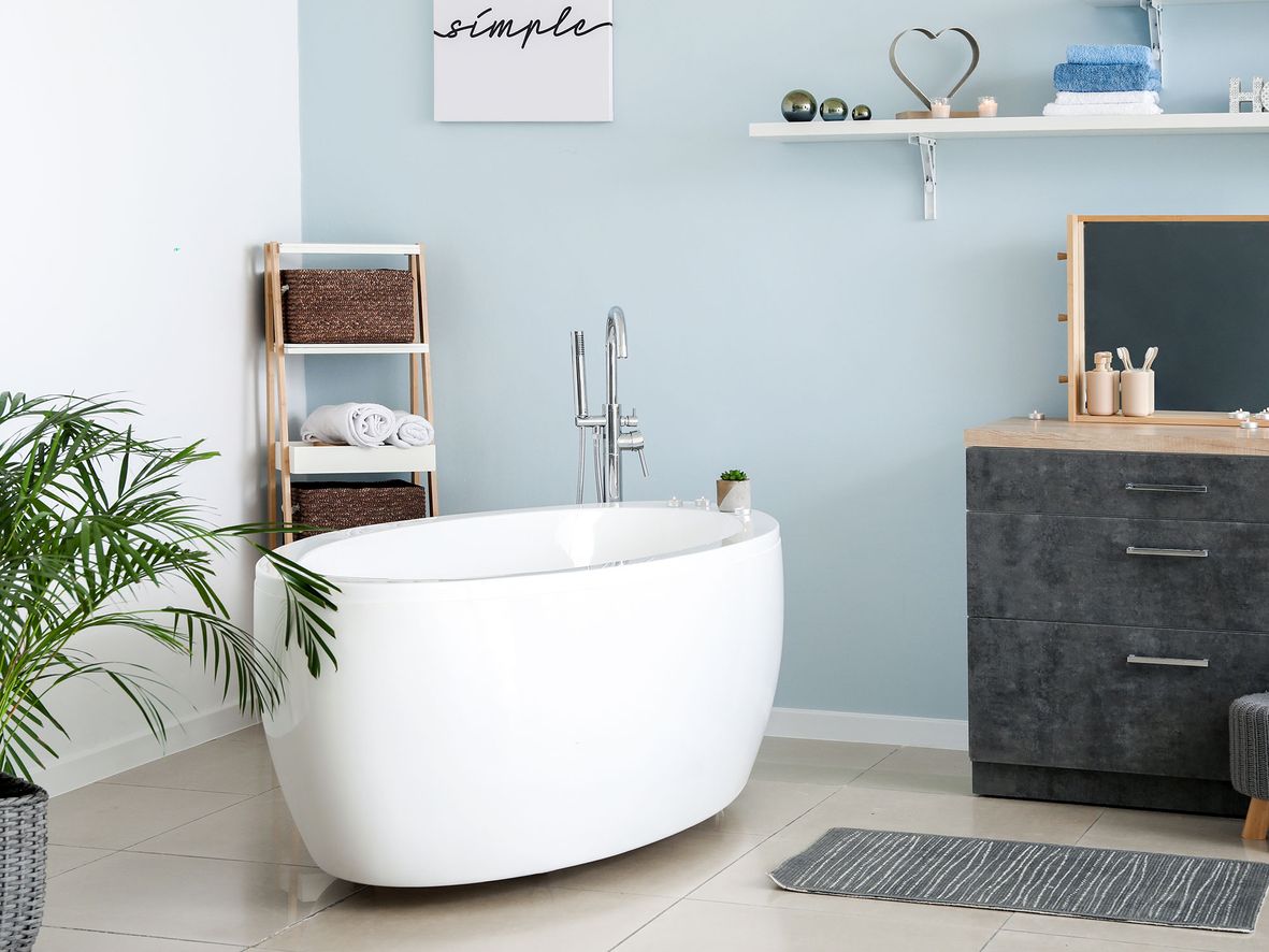 Muted bathroom with one pale blue bathroom wall, black drawer unit, white oval bath and corner shelf unit