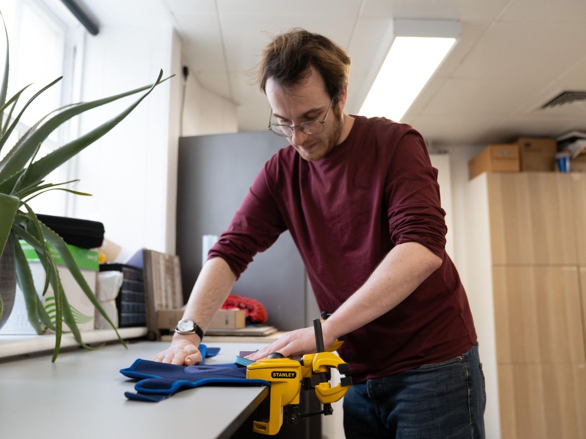 Man in maroon shirt and glasses using sandpaper on a blue knee support, held on a table by a yellow clamp