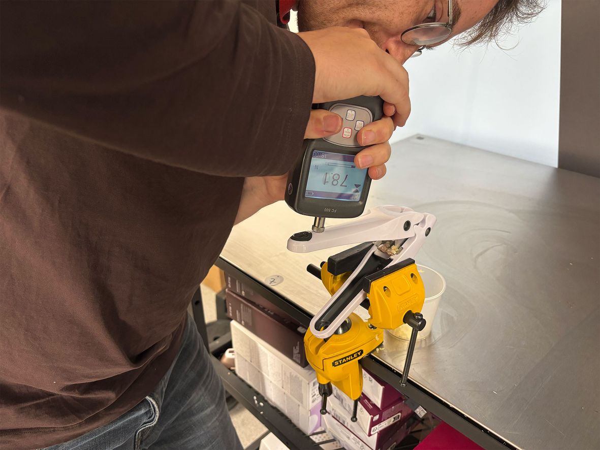 Man in brown shirt and jeans using a digital device to measure a garlic press on a metal table.