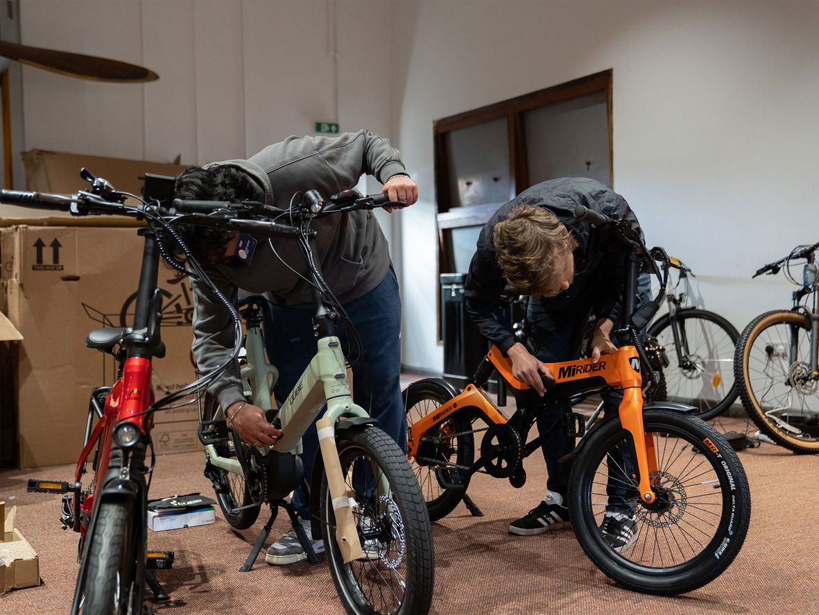 Two people working on bicycles in a workshop with tools and boxes around.