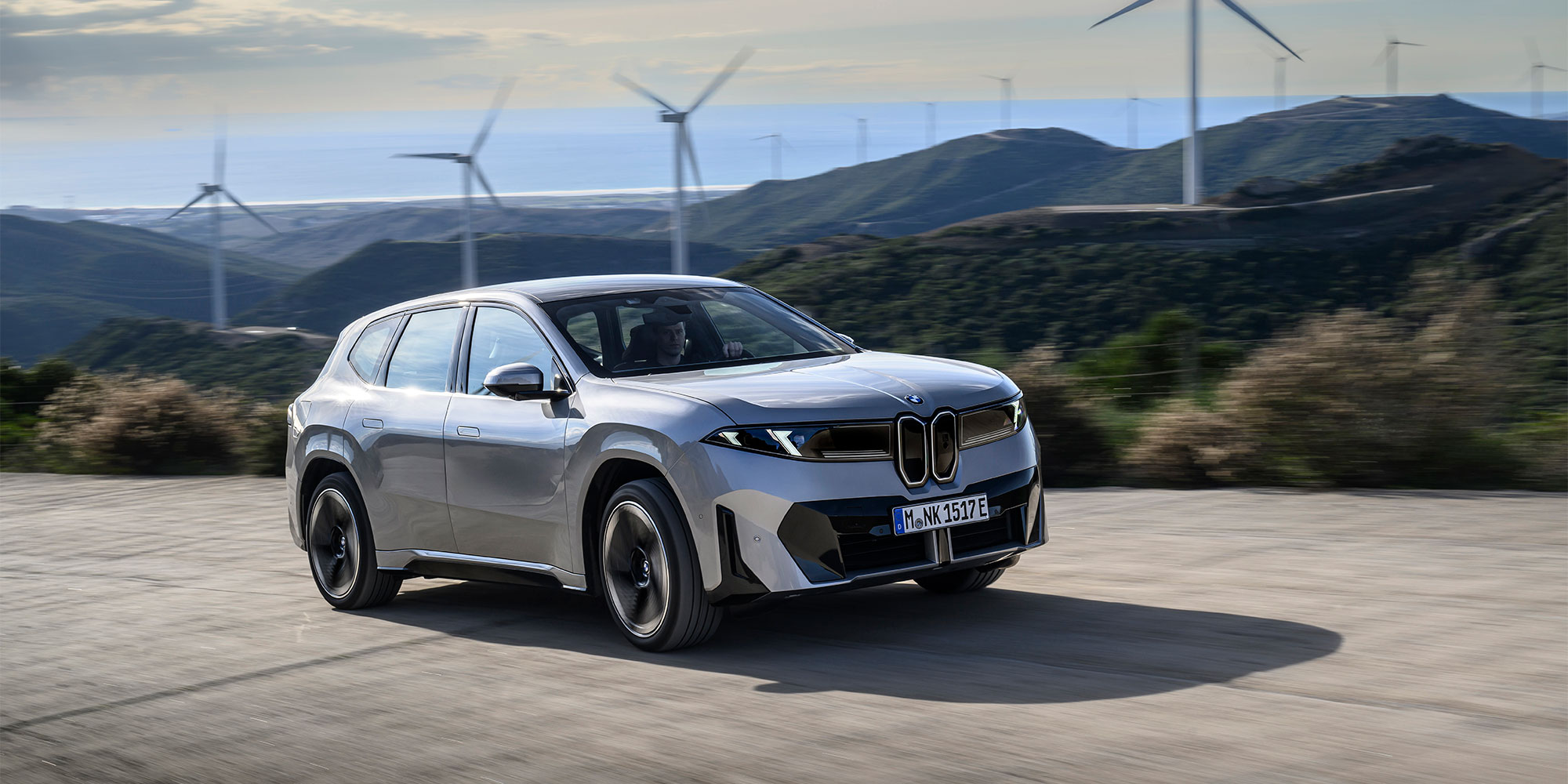 A grey BMW iX3 is driven in a landscape with wind turbines