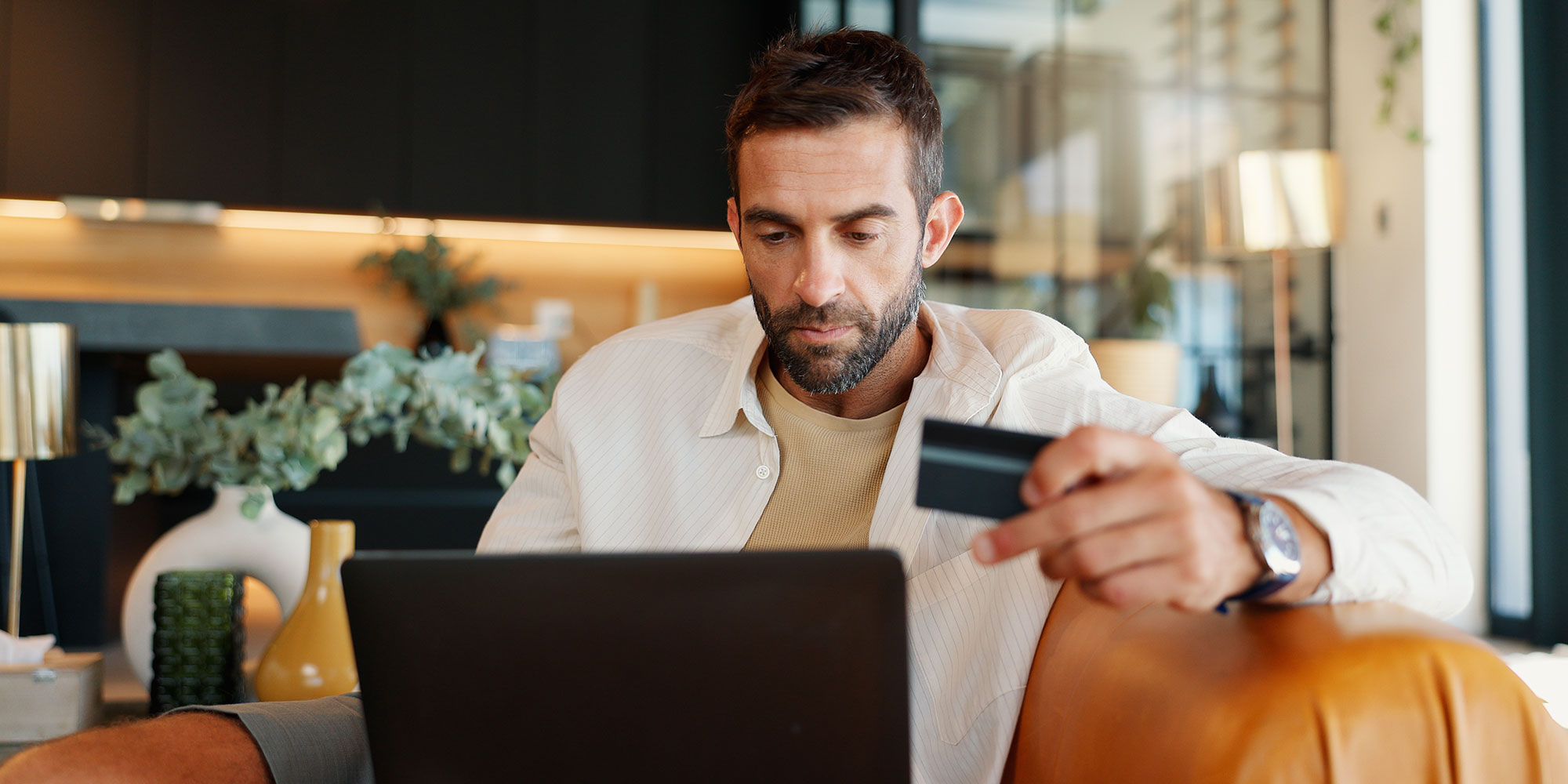 A man sitting on an orange chair with his laptop in his hand and his bank card in his left hand