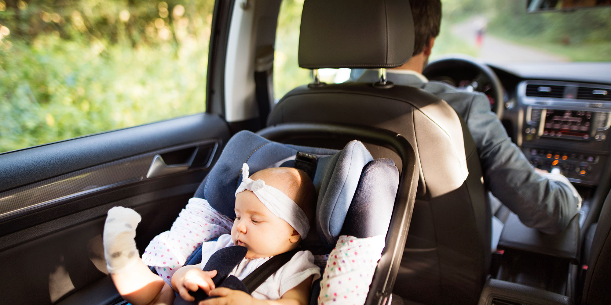 An infant wearing a white headband lies in a rear-facing safety seat in the back of a car while a man drives.