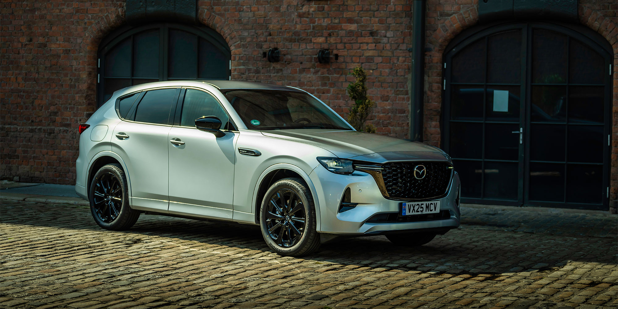 A grey Mazda CX-60 PHEV, parked on cobbles near a dock building.