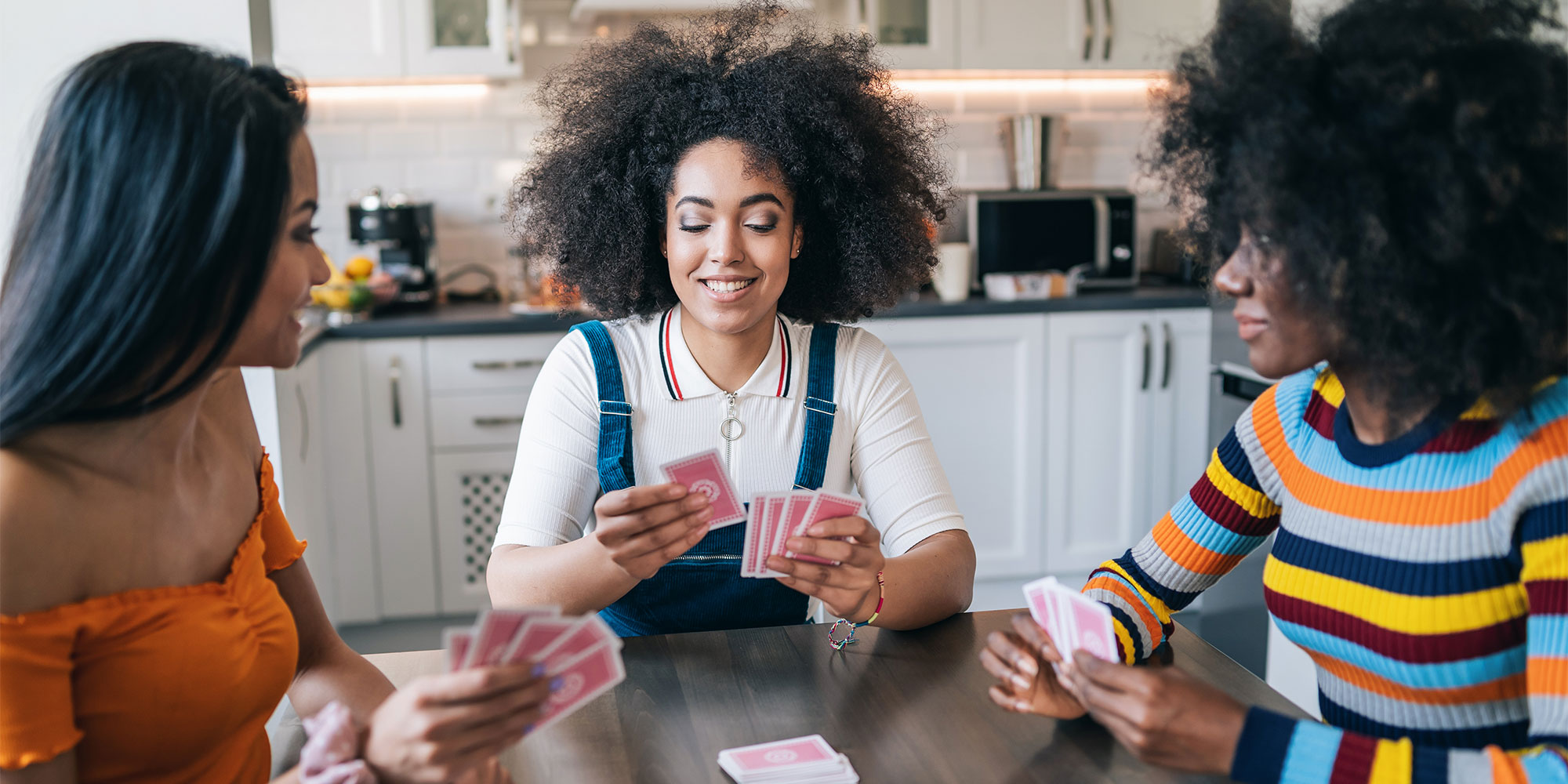 Three women playing a card game in a kitchen. One woman is smiling.