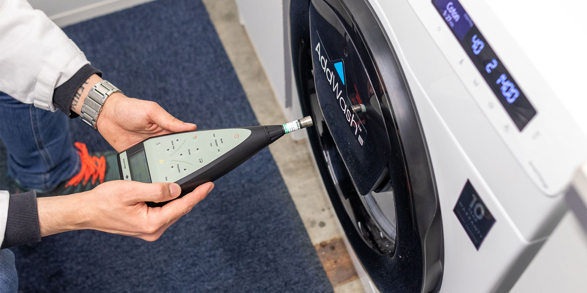A lab technician monitors a washing machine's normal operating noises using a decibel monitoring device