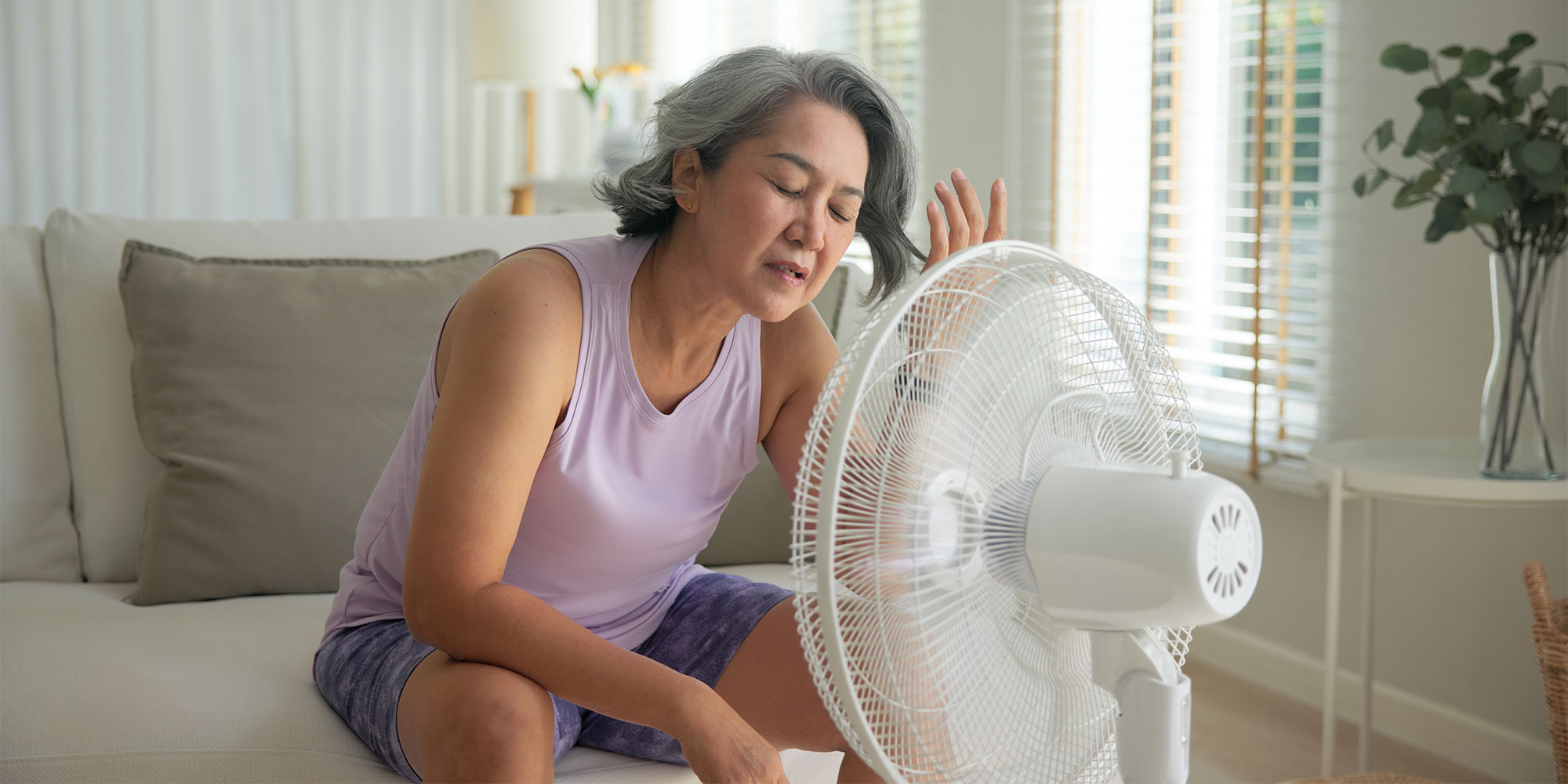 Woman in purple tank top and gray shorts sitting on couch with white fan.