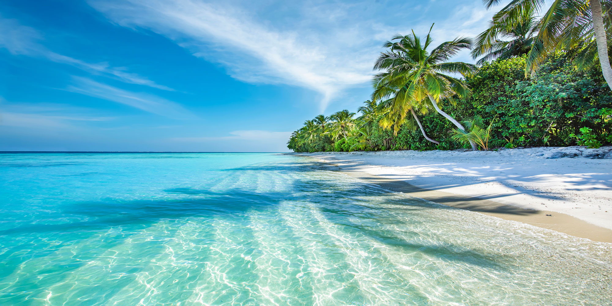 Clear blue ocean waves gently lapping onto a pristine white sandy beach lined with lush green palm trees and dense vegetation under a partly cloudy sky.