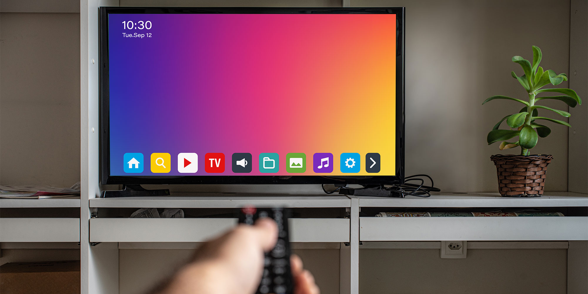 A blurred hand holds a TV remote pointing at a TV. The screen shows a series off app icons