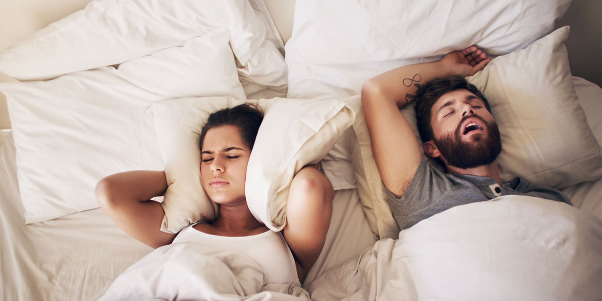 Woman covering her ears with pillow as man beside her snores