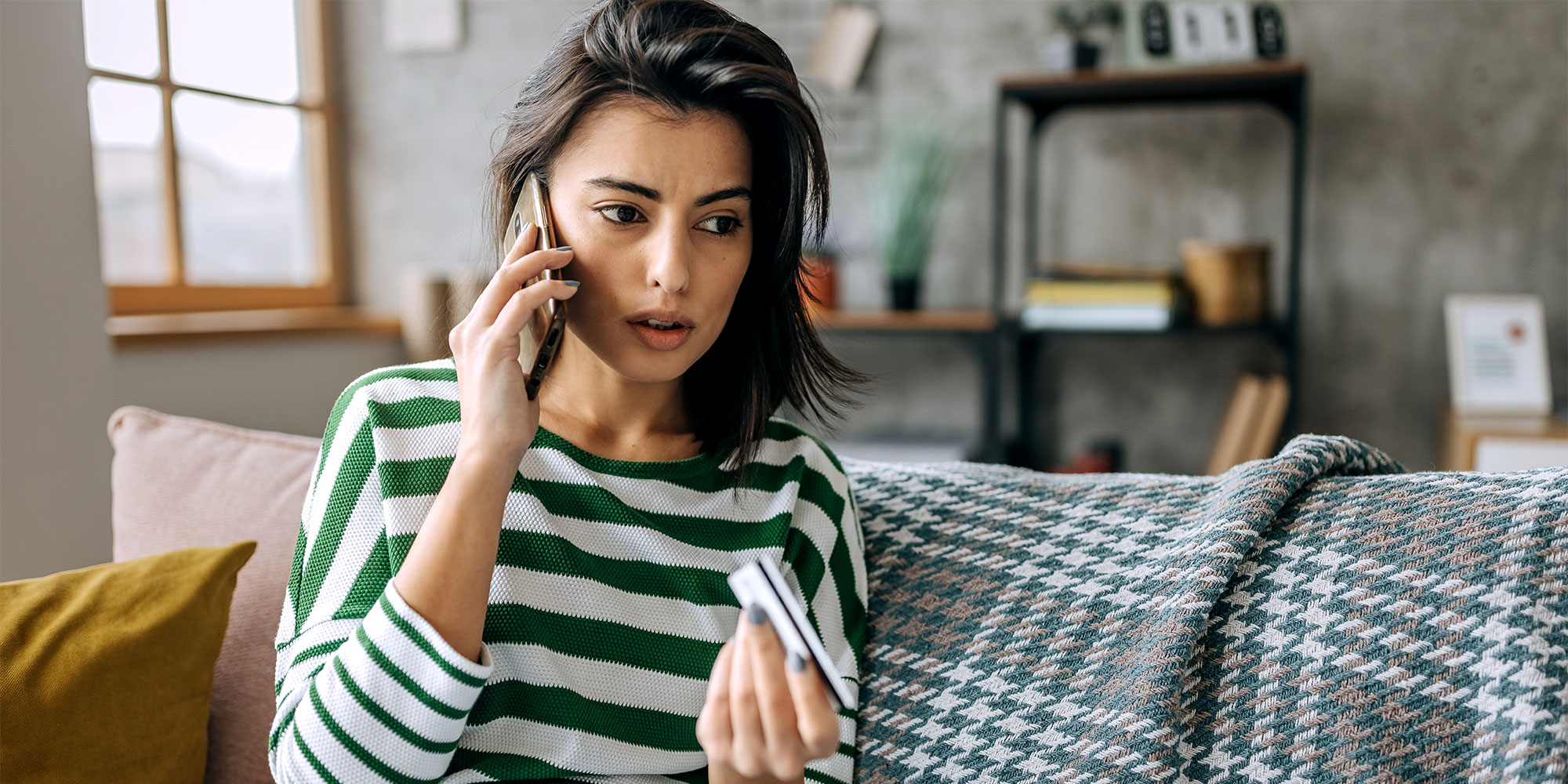 A woman holds a mobile phone to her ear and holds a bank card in her other hand, while looking anxious.