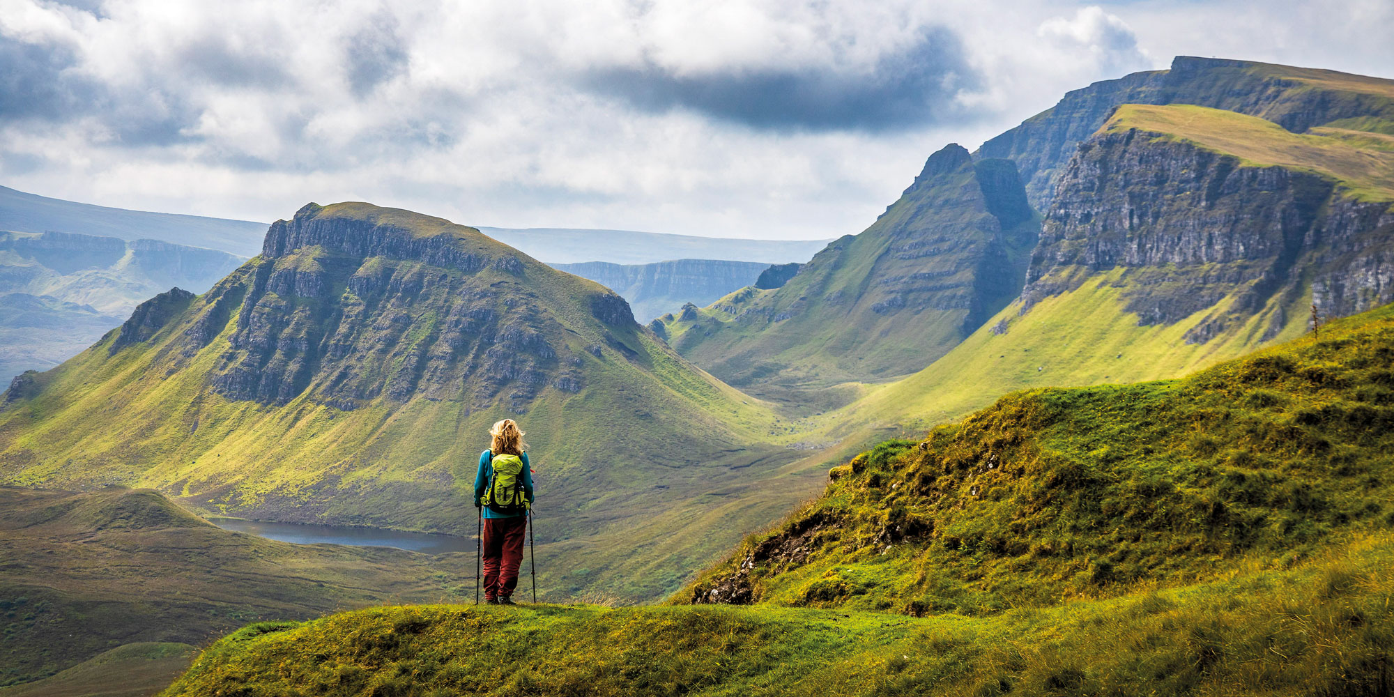 Hiker with backpack and trekking poles stands on grassy mountain ridge overlooking valley and lake.