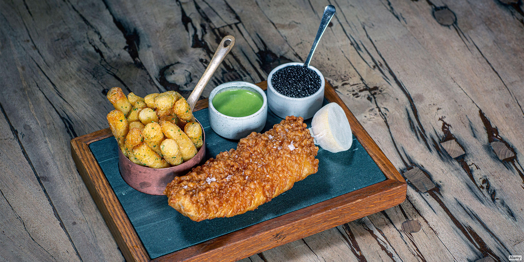 Wooden tray with fish, fries, green sauce, black caviar, and white sauce on a wooden table.