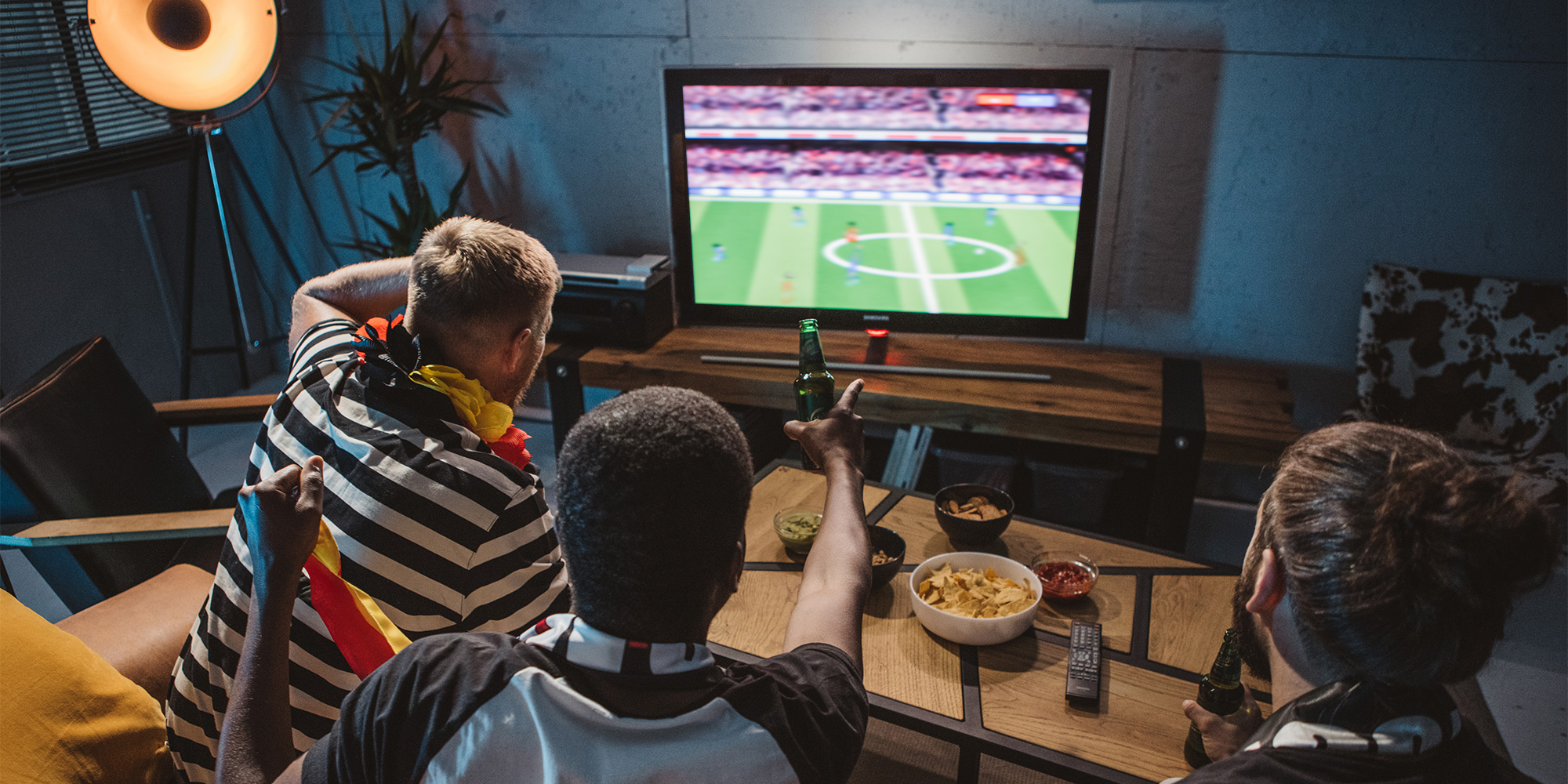 Three men are watching a football game in their living room. They are drinking beer and eating crisps