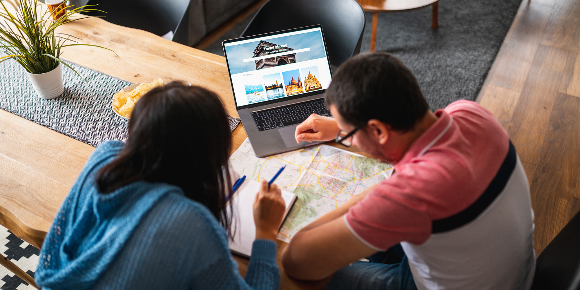A couple plans a holiday on a laptop at home on a coffee table, with a notepad, pens and and map.