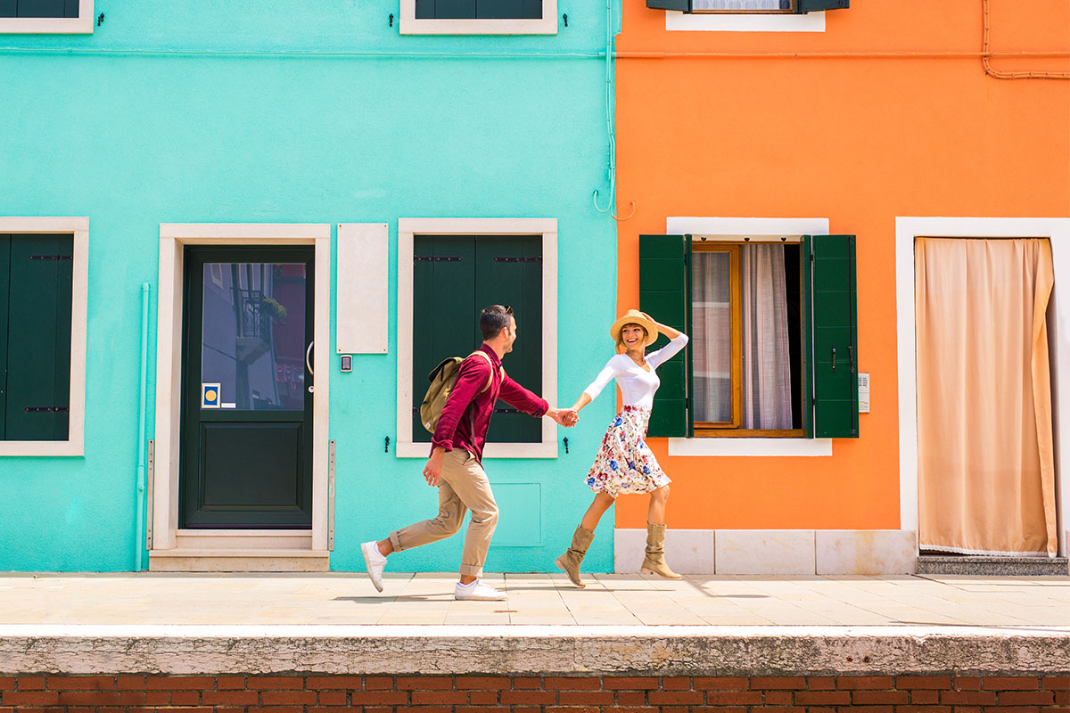 Couple holding hands, running on sidewalk past colorful buildings. Man in red shirt, woman in white top and floral skirt.