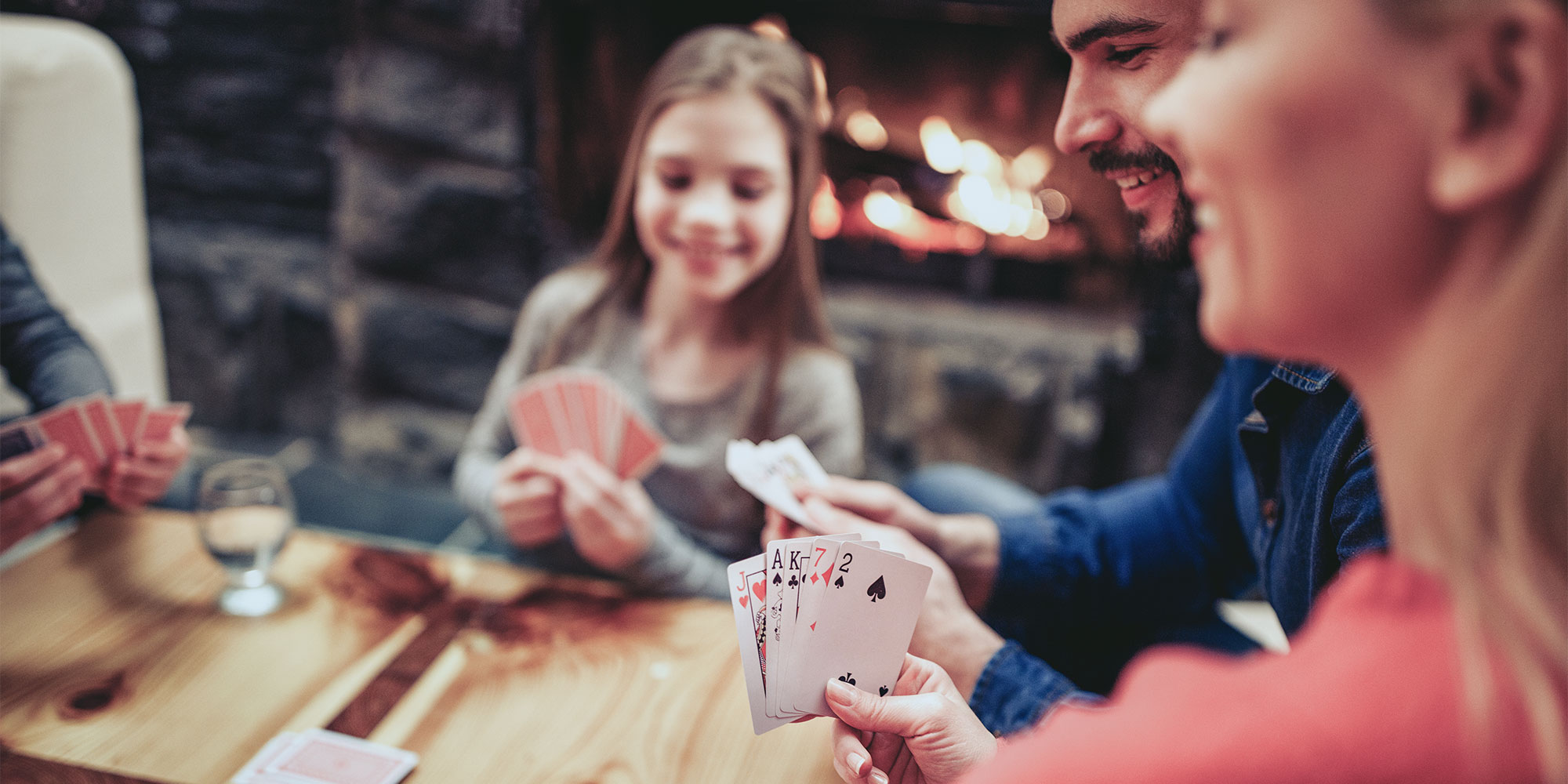 Group of people playing cards around a table in front of a fireplace.