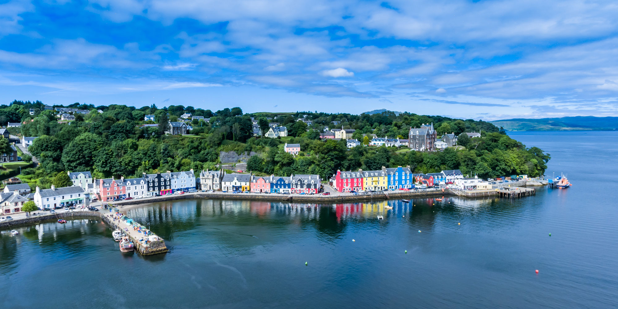 Aerial view of coastal Mull with colorful houses, boats, and a harbor under a blue sky.