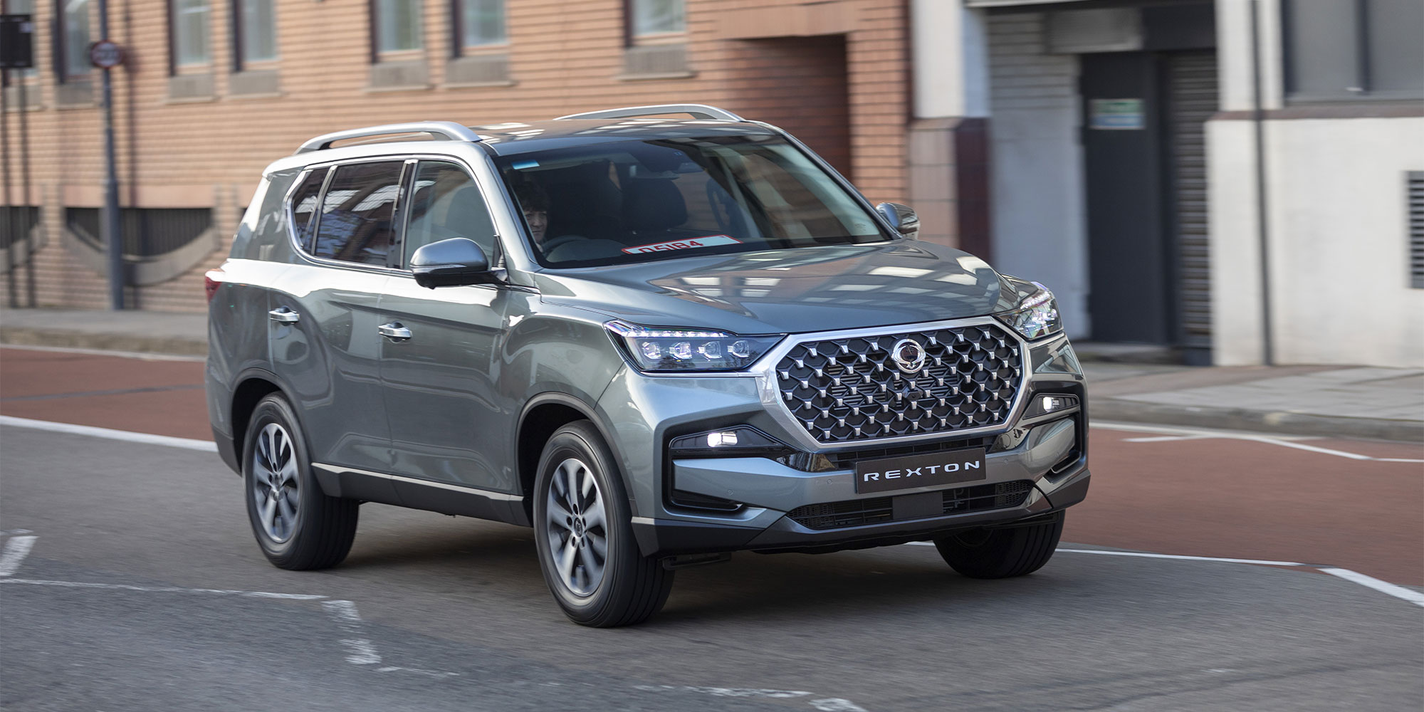 Grey KGM Rexton SUV driving on a city street with buildings in the background.
