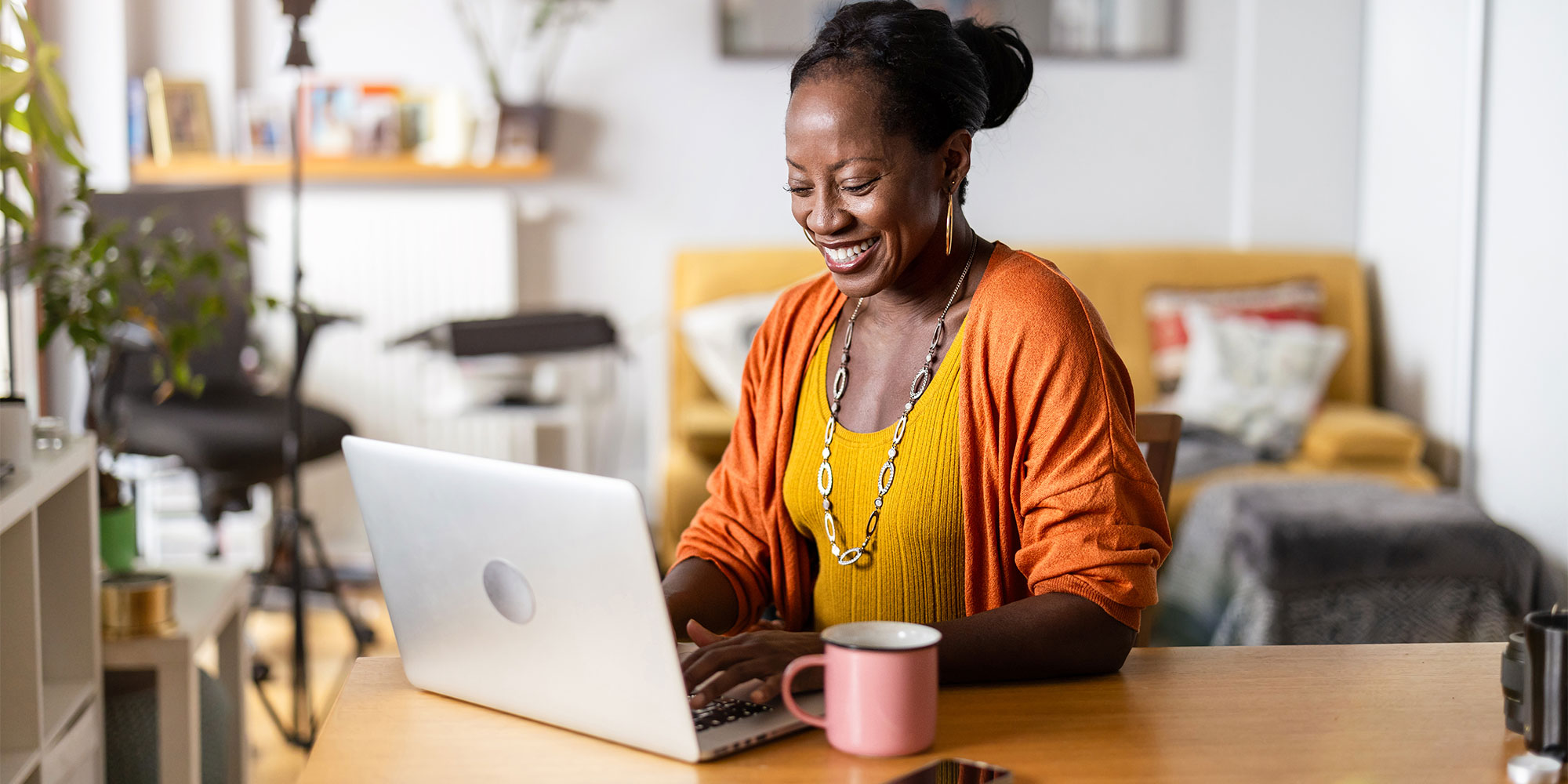 Woman in orange cardigan and yellow top smiling while working on laptop at home.