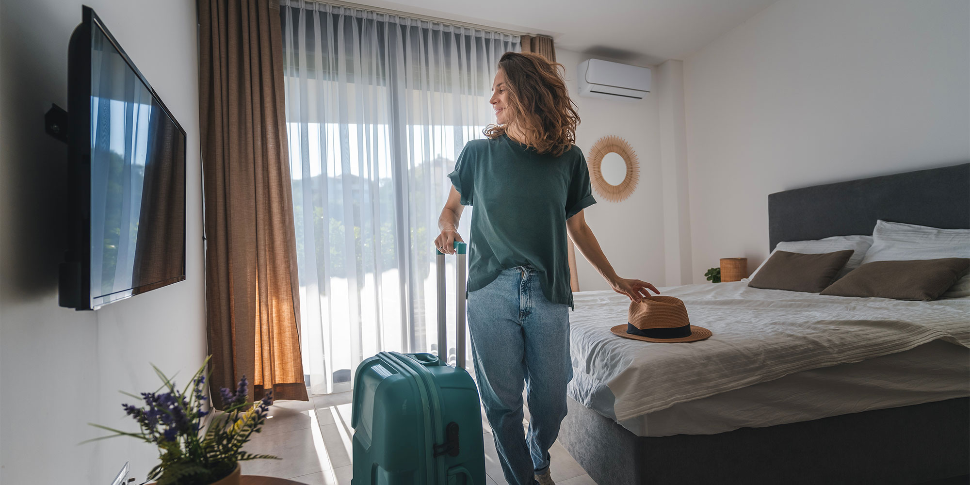 Woman in green shirt and jeans pulling teal suitcase in hotel room with bed and TV.