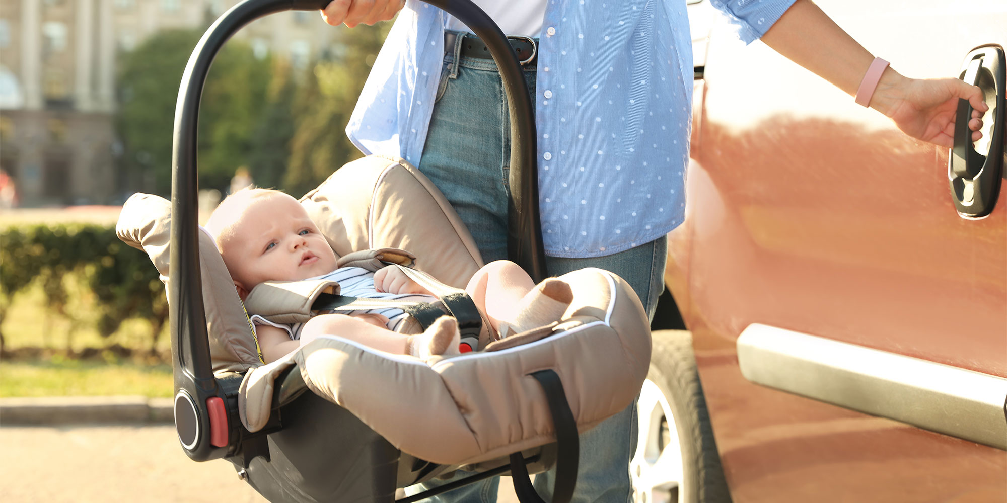 A person carries an infant car seat with a baby inside toward an orange car. One hand is holding the car seat handle while the other reaches for the car door.