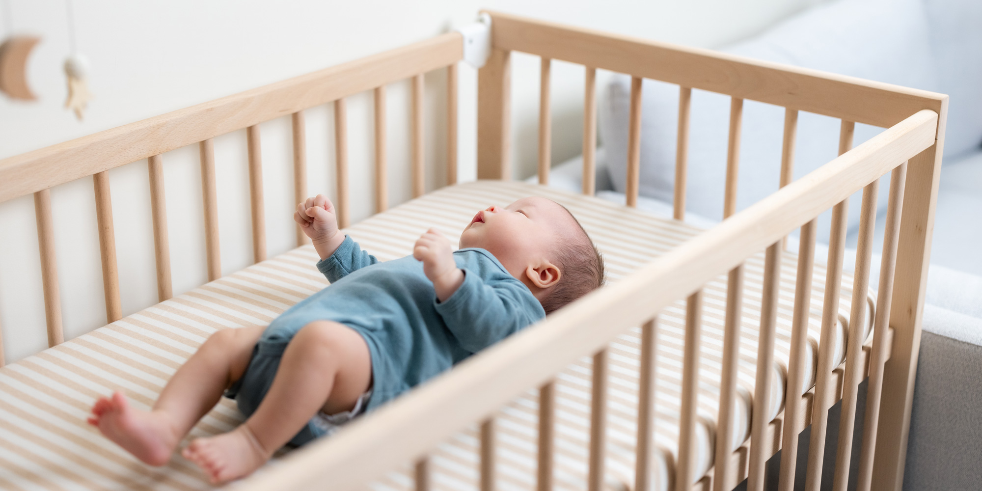 A high-angle, side view of a baby in a blue onesie lying on their back in a wooden cot. The cot has a striped mattress protector, and a celestial-themed mobile with a wooden moon and stars hangs partially in view above.