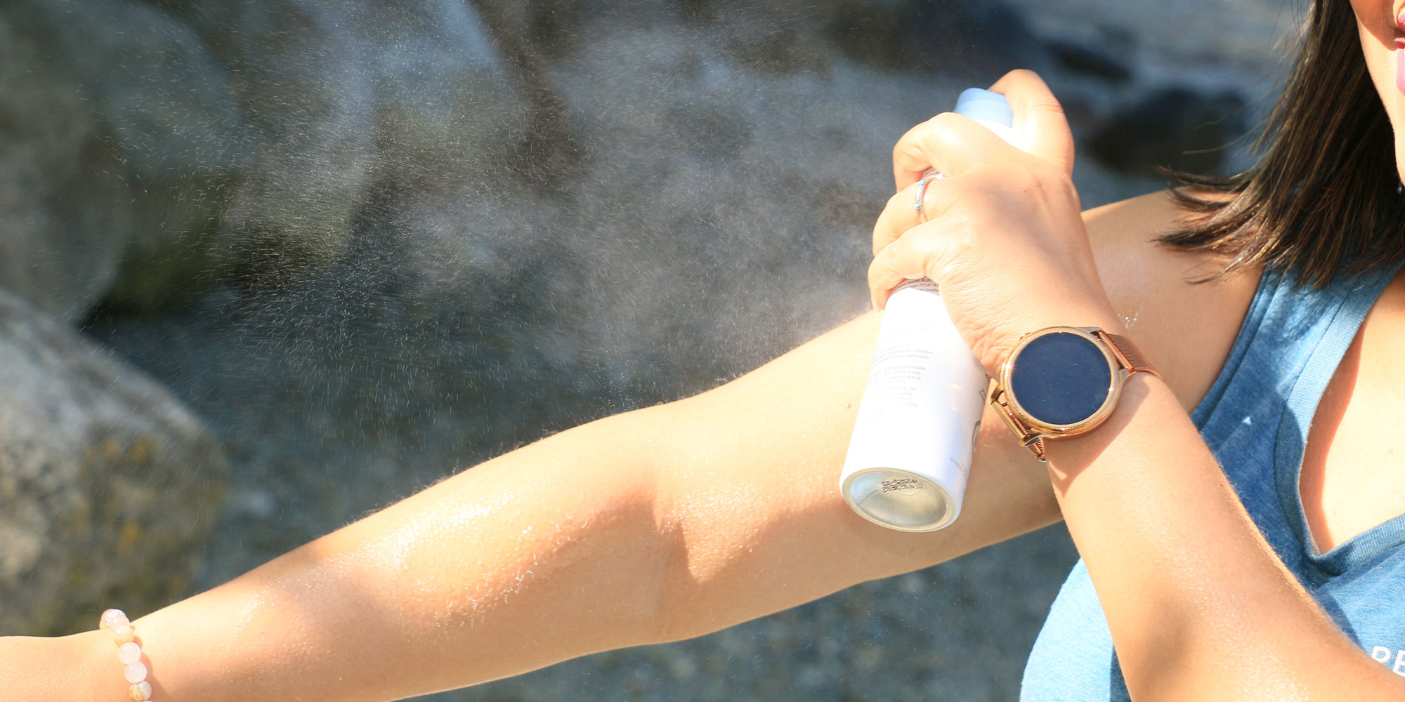 A person spraying a fine mist of aerosol sunscreen onto their arm outdoors, with a blurry rocky background