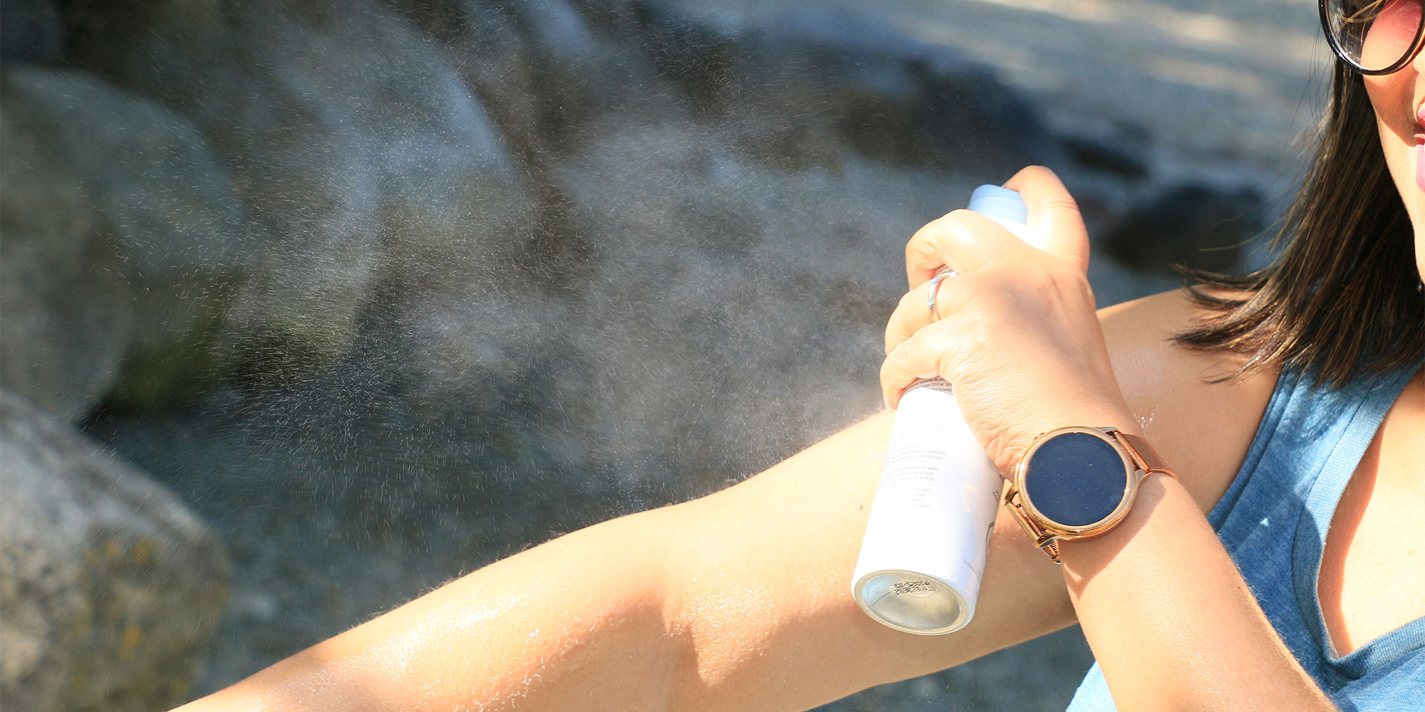 Close-up of a woman in a blue tank top and smartwatch spraying a fine mist of aerosol sunscreen onto her extended forearm outdoors.