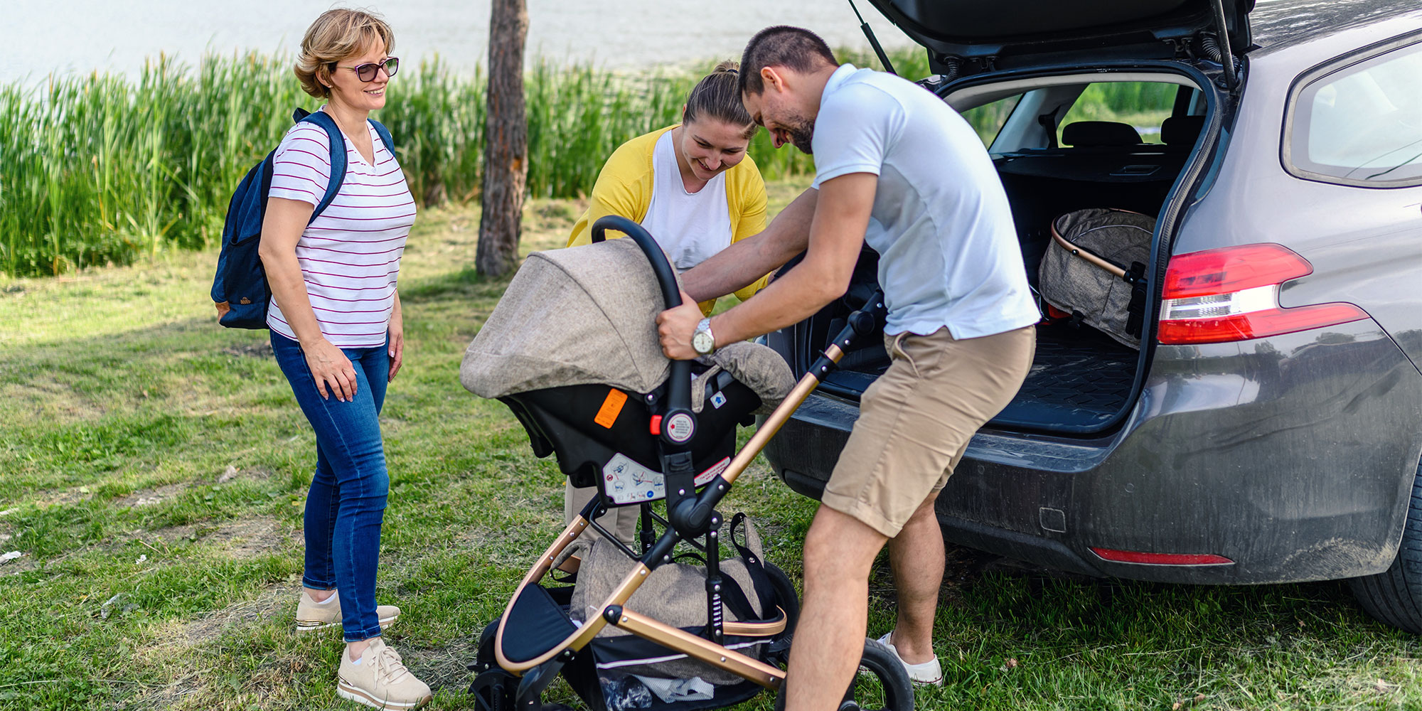 A group of three adults stands behind a gray car with its trunk open. A man is adjusting an infant car seat onto a stroller frame while two women watch and smile.