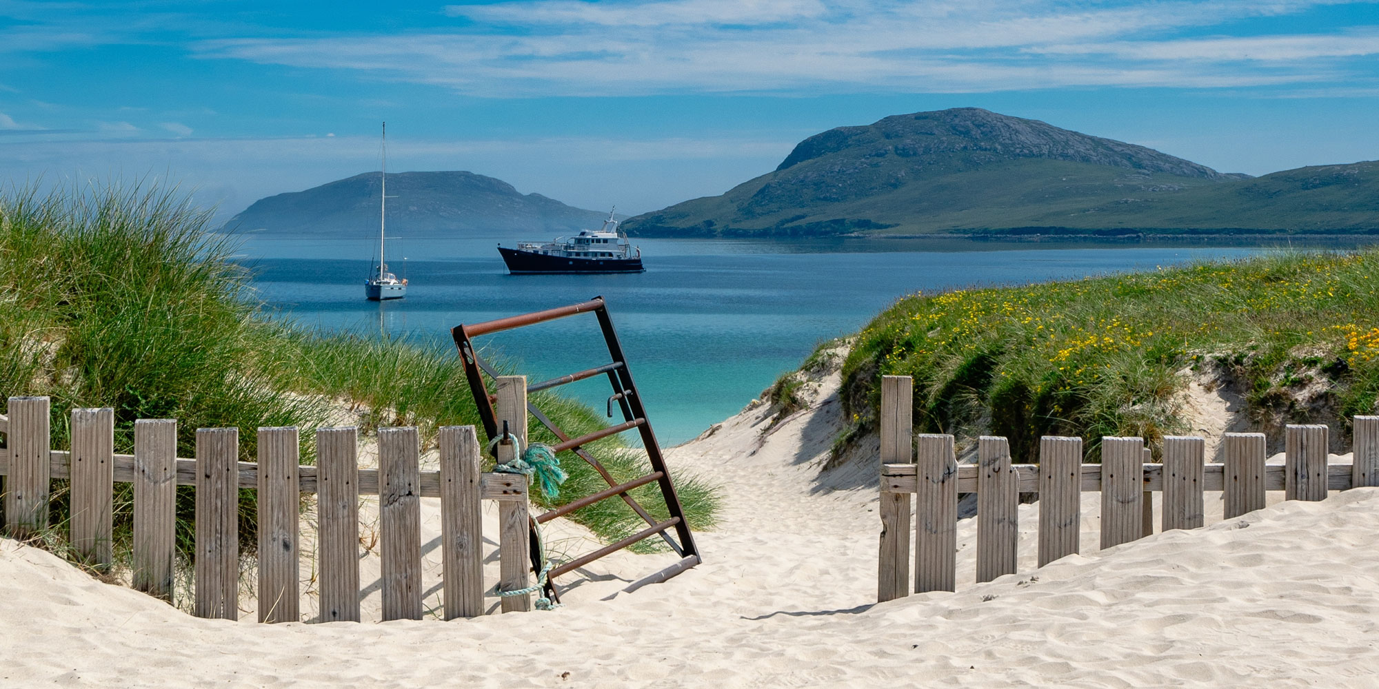 Wooden fence on sandy beach with boats in water and mountains in background on the Isle of Barra