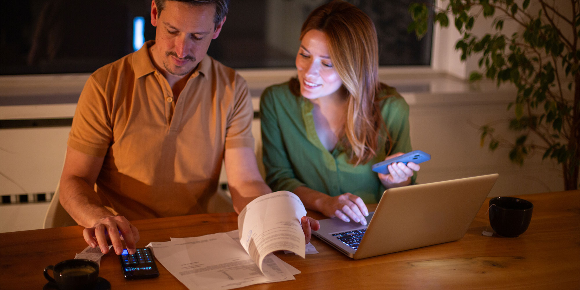 A smiling young couple sit at a desk with a laptop, calculator and paperwork in front of them.