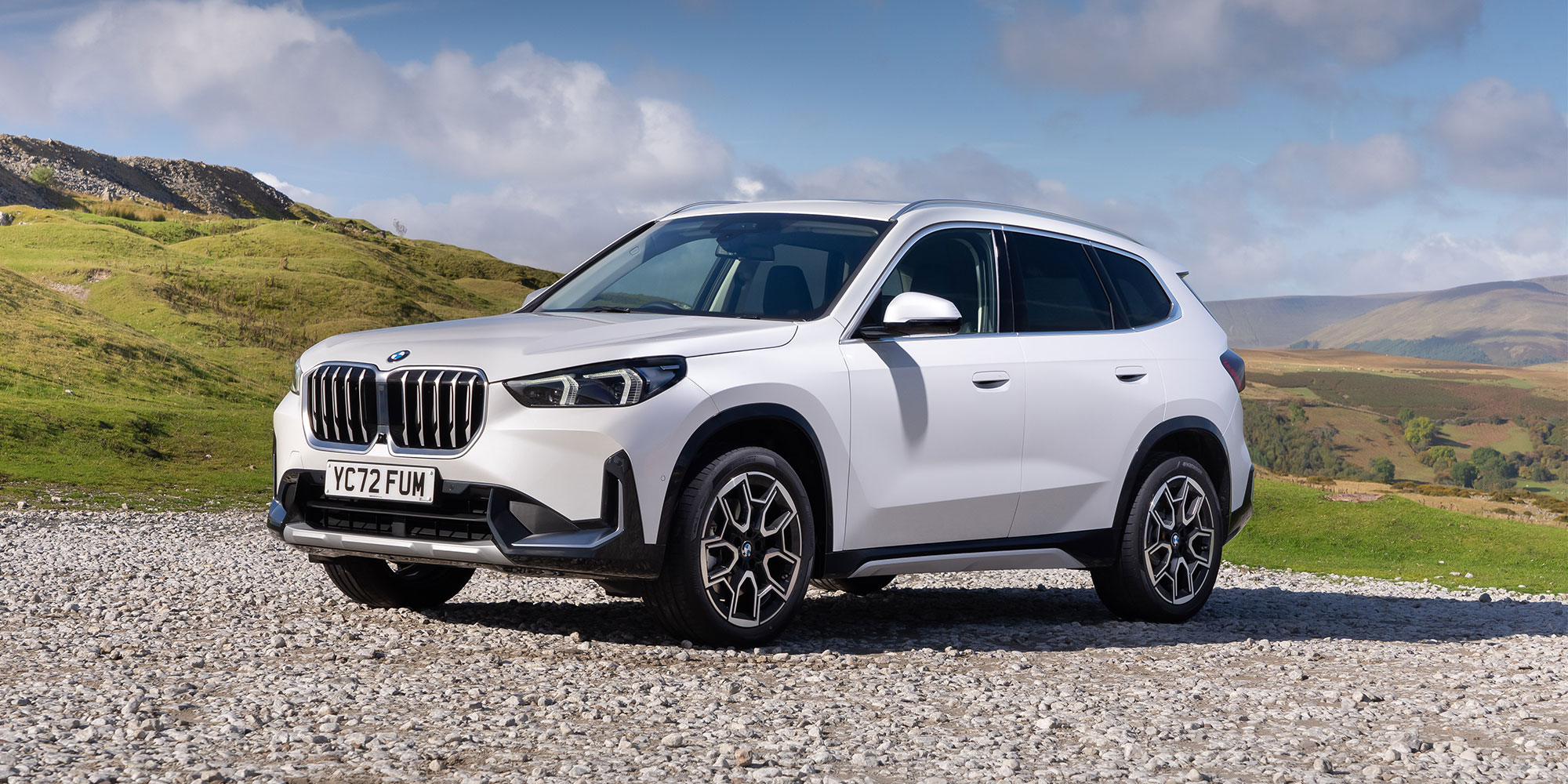 A white BMW X1 parked on gravel in front of a hill.