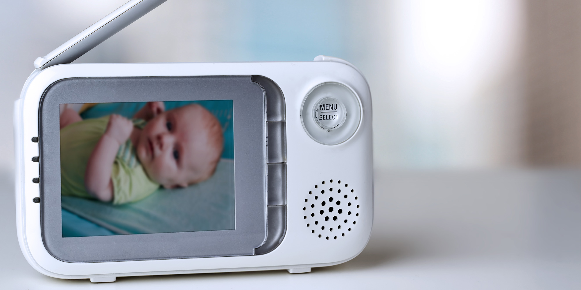 A close-up of a white digital video baby monitor sitting on a light-colored surface. The monitor's screen shows a live, slightly blurred feed of a baby lying down. The device features a "Menu/Select" button, a small speaker grille, and an antenna extended at the top.