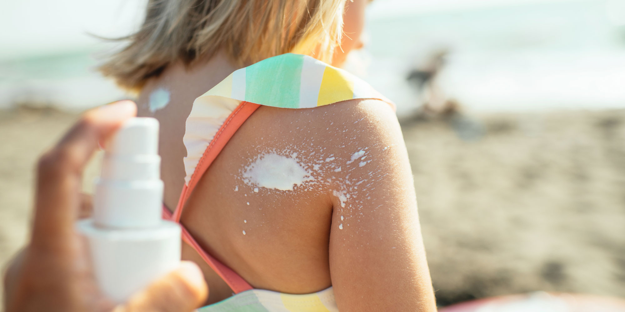 A hand holding a white bottle and spraying sun cream onto a child's shoulder at a sunny beach