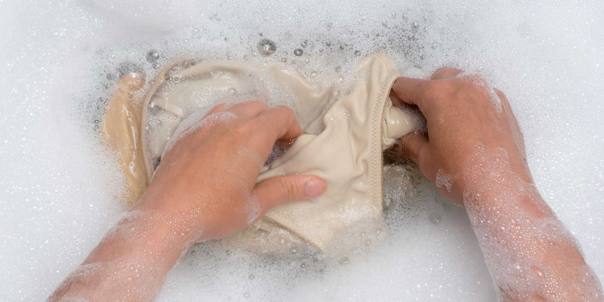 A person's hands are washing a pair of white pants in a tub of soapy water.