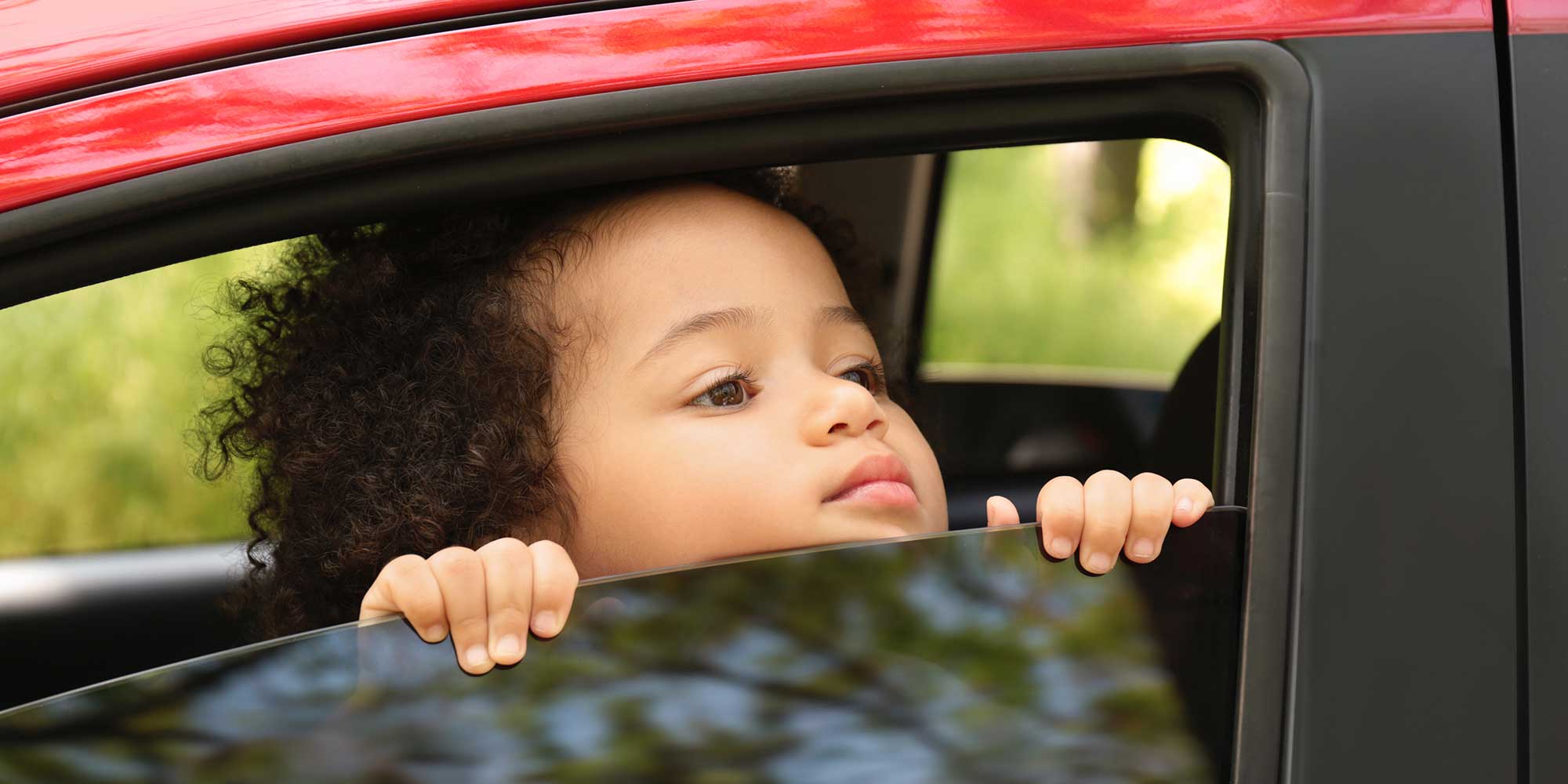 A young child with curly dark hair looking out the open window of a red car, resting their hands on the top edge of the glass.