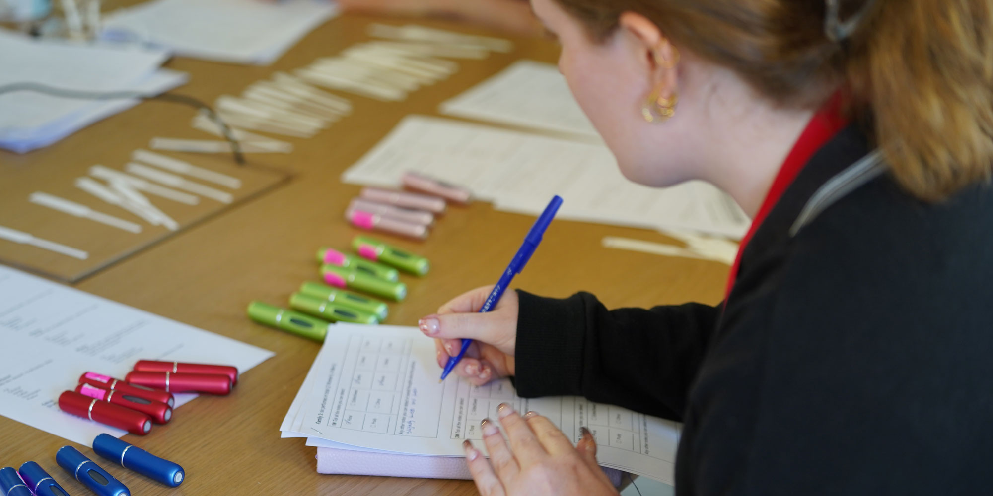 Which? perfume tester writing notes on white paper, surrounded by blue, red, green and pink atomiser spray bottles