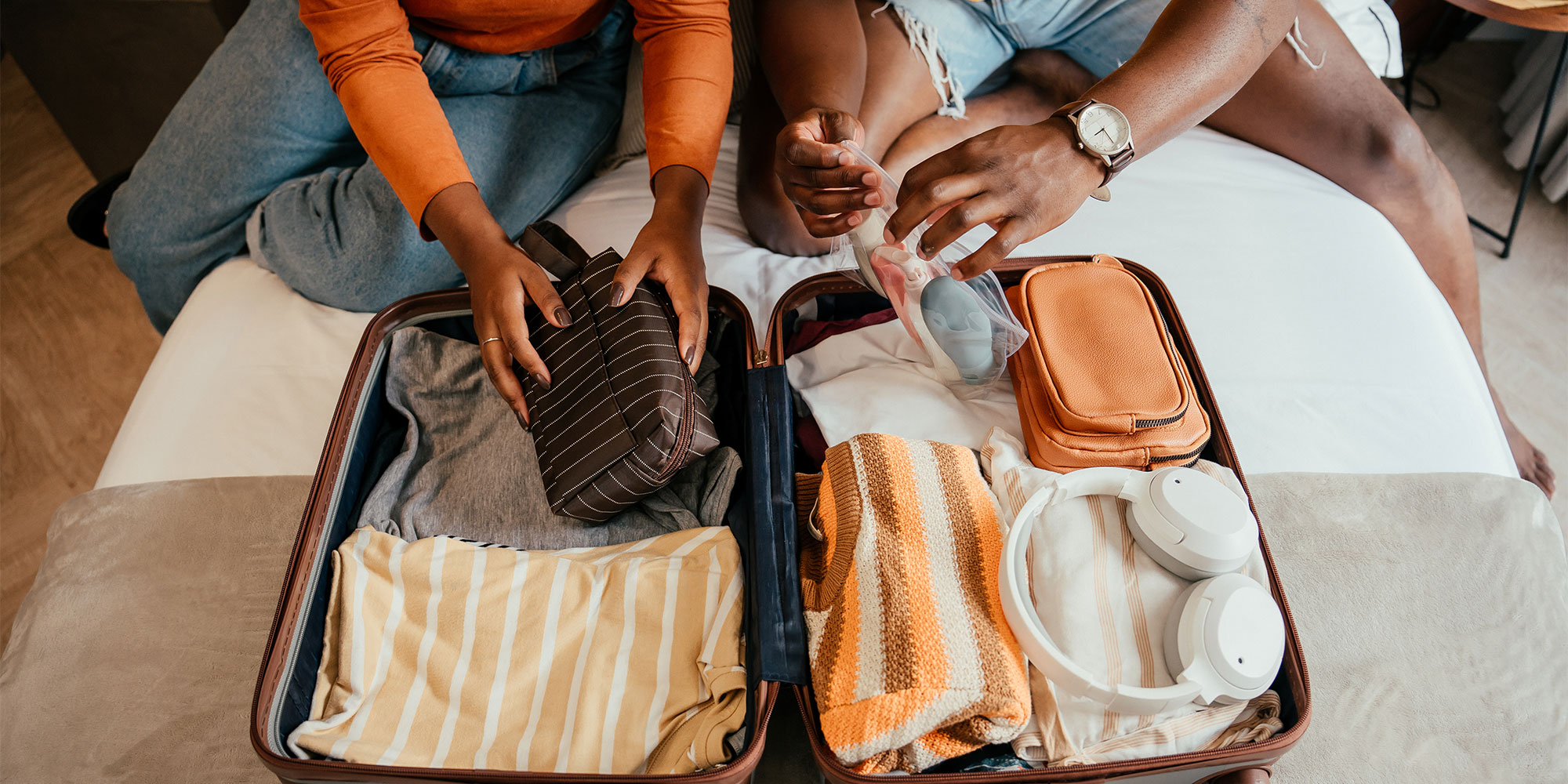 Couple putting wash bag into an open suitcase that is filled with items including clothes and white headphones