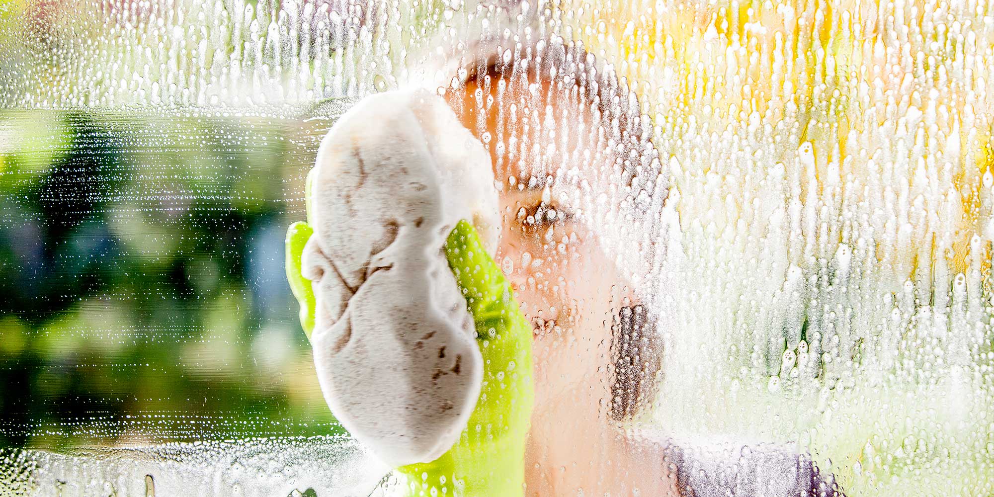 A window covered in bubbles being wiped with a cloth by someone wearing yellow washing-up gloves.