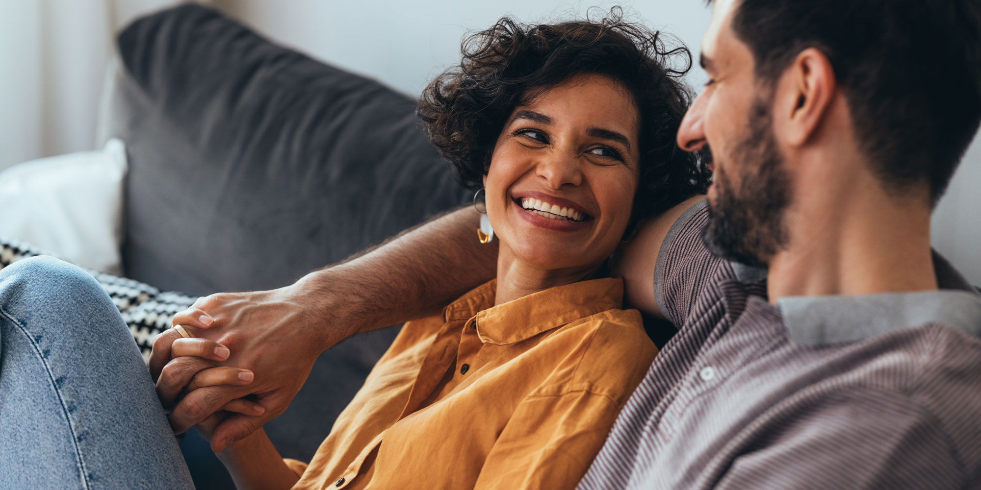 Women in yellow top smiling and holding a man's hand while they are sat on a grey couch