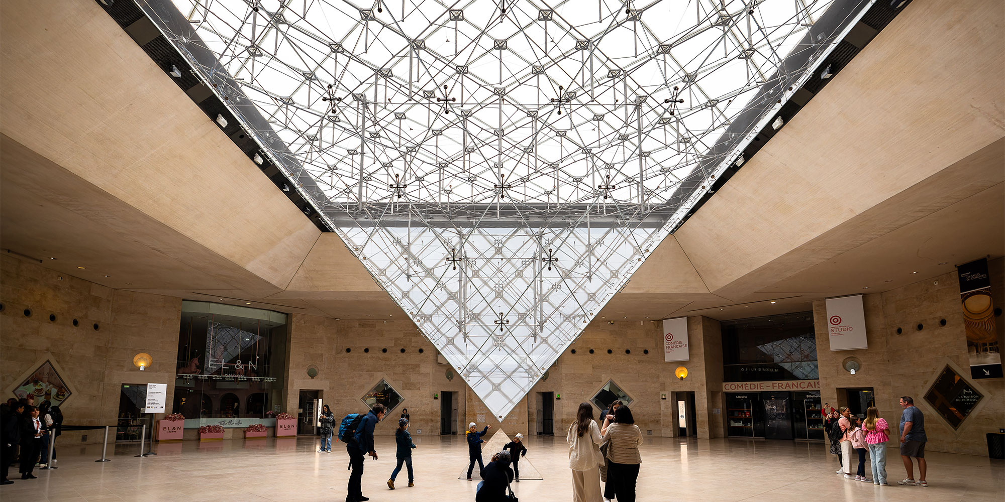 People gather and explore beneath a large, geometric glass pyramid in a spacious, light-filled lobby.