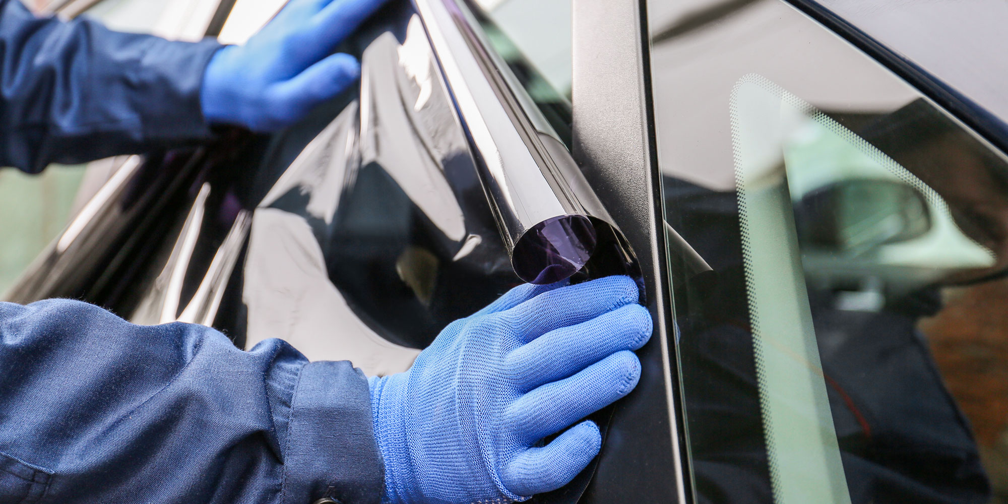 Person in blue jacket and blue gloves applying window tint to a car.
