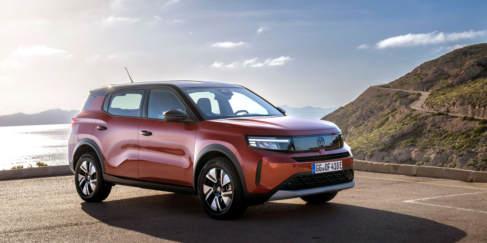 A red Vauxhall Frontera is parked on tarmac near a coastline and a hill road
