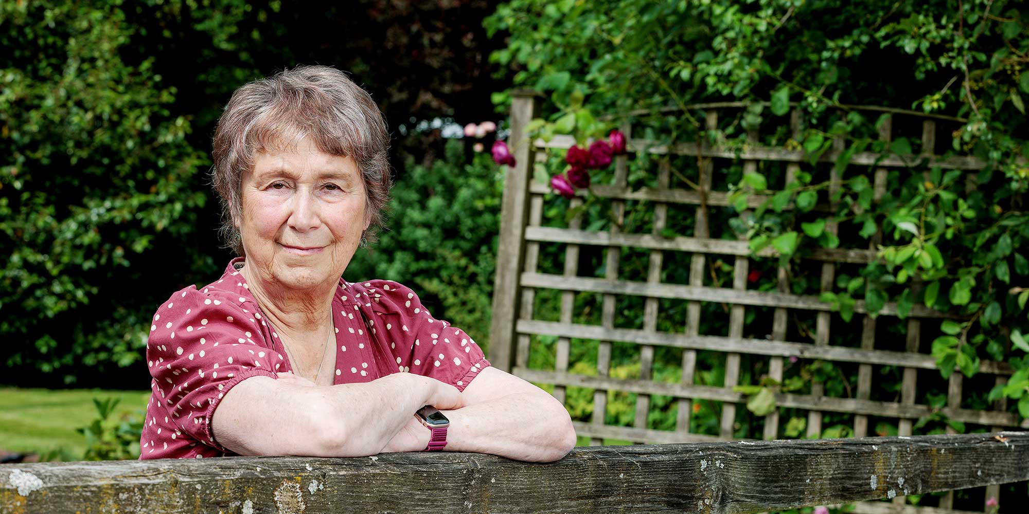A woman in her 80s with short hair is leaning against a fence smiling