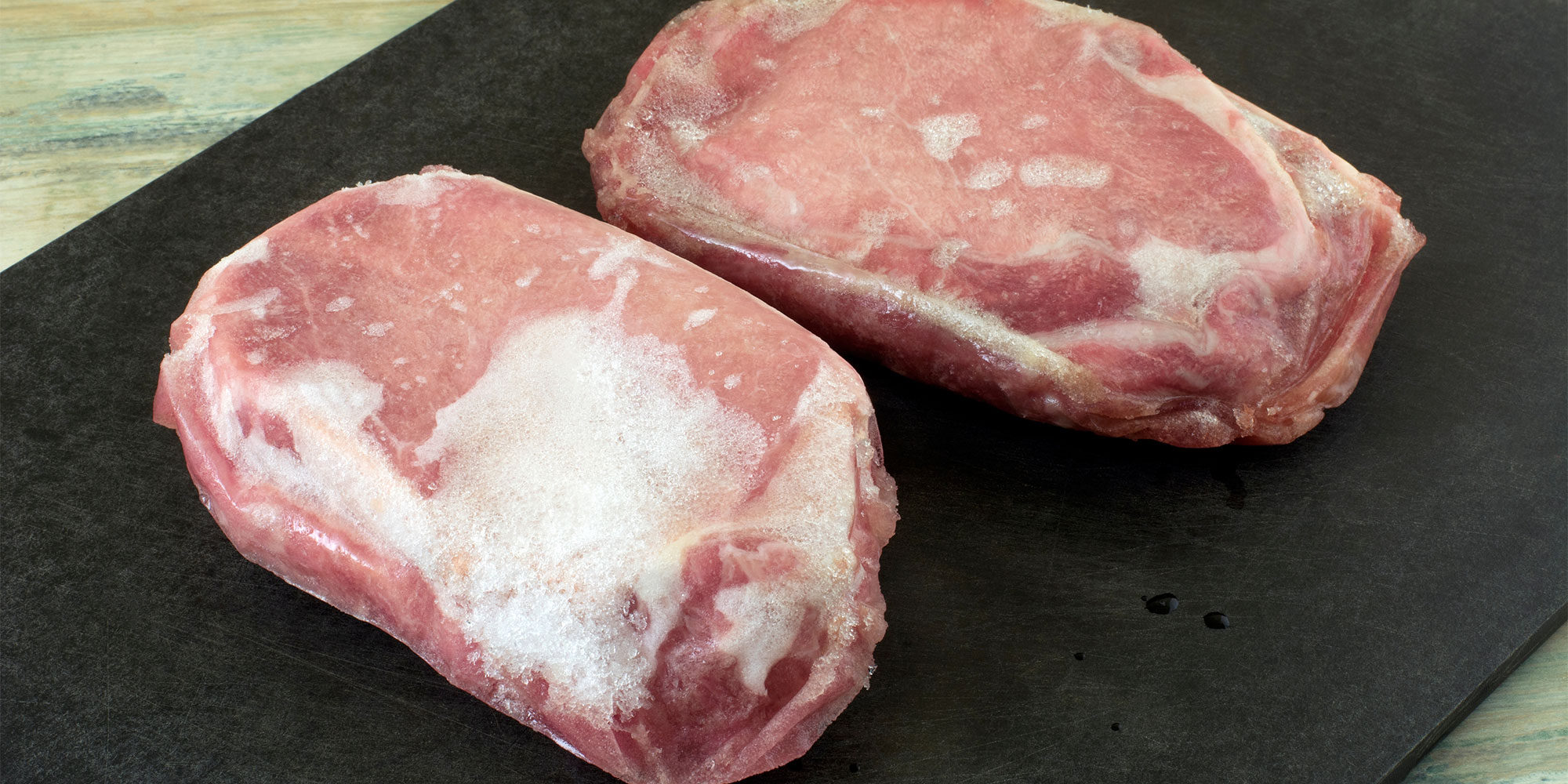Two raw pork chops on a black chopping board. Both cuts of meat show significant freezer burn, characterised by large, dry-looking white patches and crystallized frost on the surface.