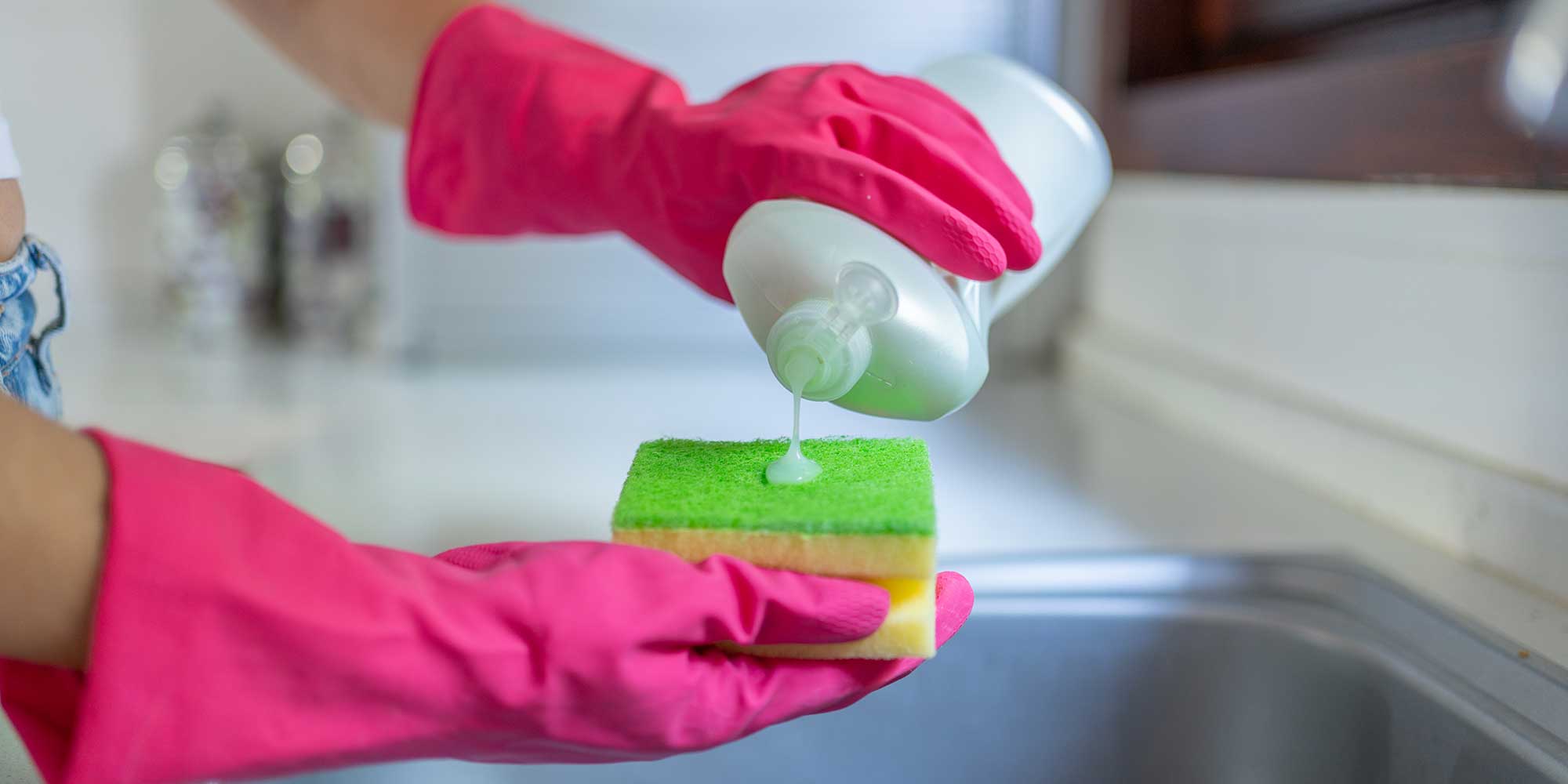 An unbranded bottle of washing-up liquid being squeezed onto a yellow and green sponge by someone wearing pink washing-up gloves.