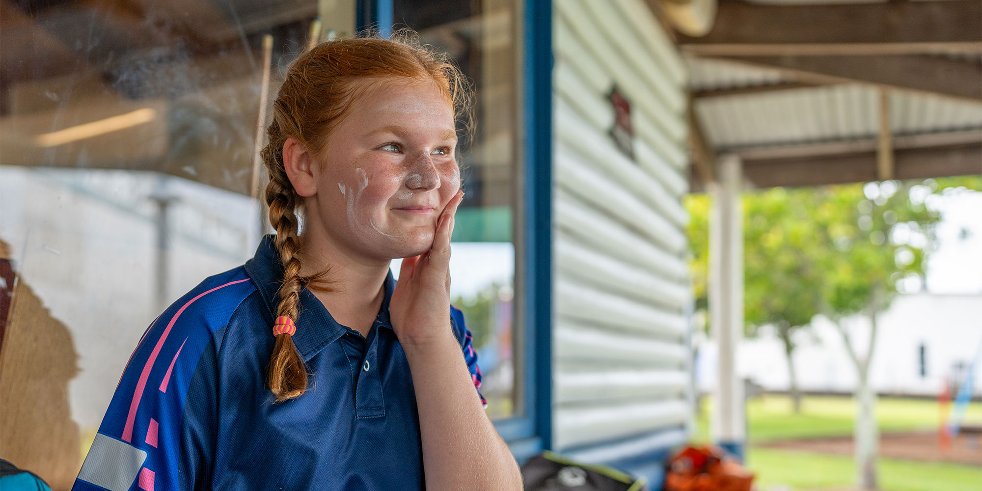 A young red-haired girl with braided hair and a blue polo shirt smiling as she rubs white sunscreen lotion into her cheek under an outdoor pavilion.