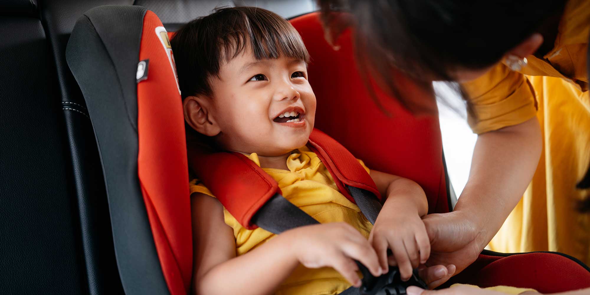 A cheerful young toddler in a yellow shirt smiling as an adult helps buckle them into a bright red car seat harness.