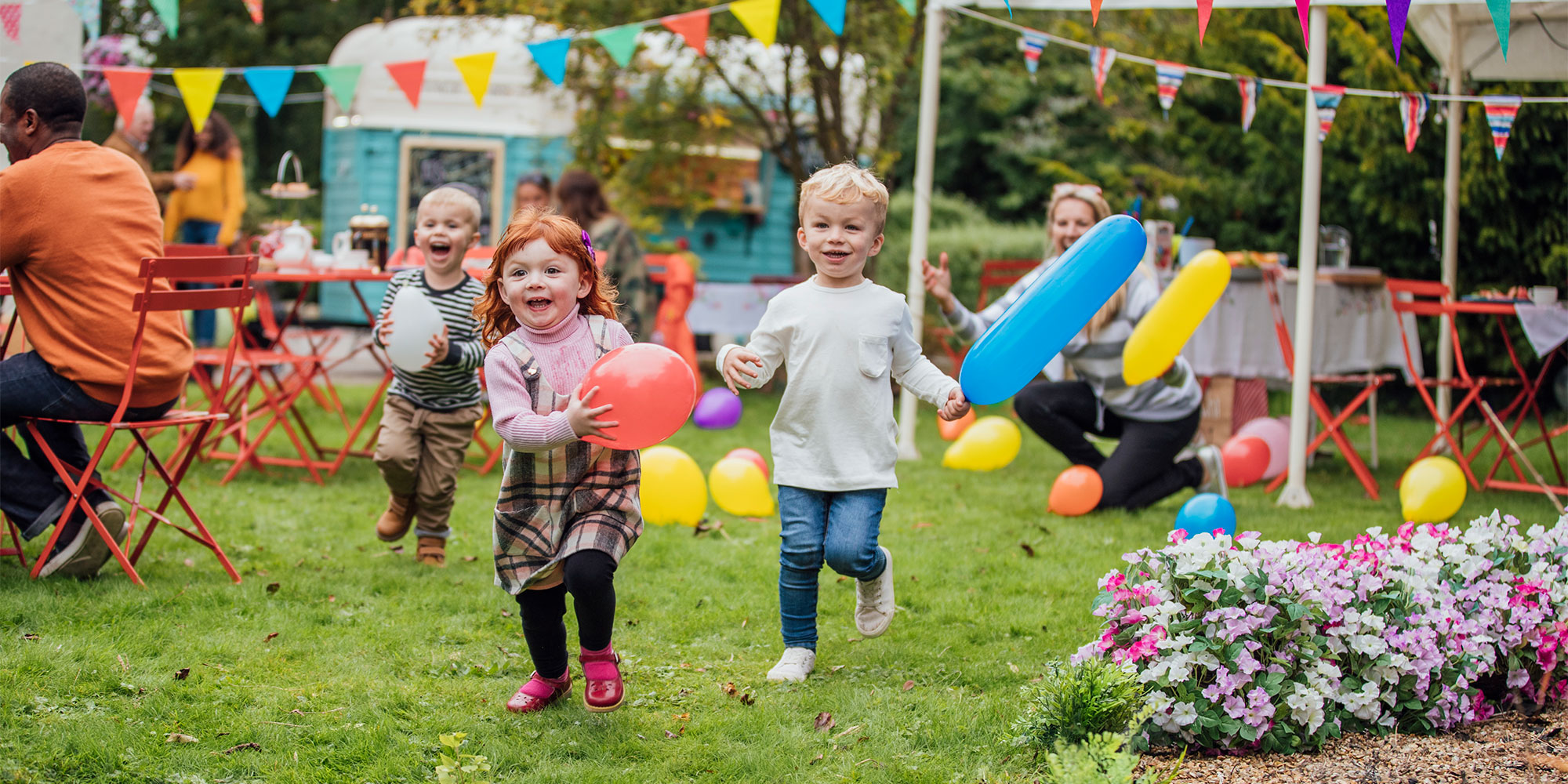 Three children running and holding balloons with a woman watching on from behind